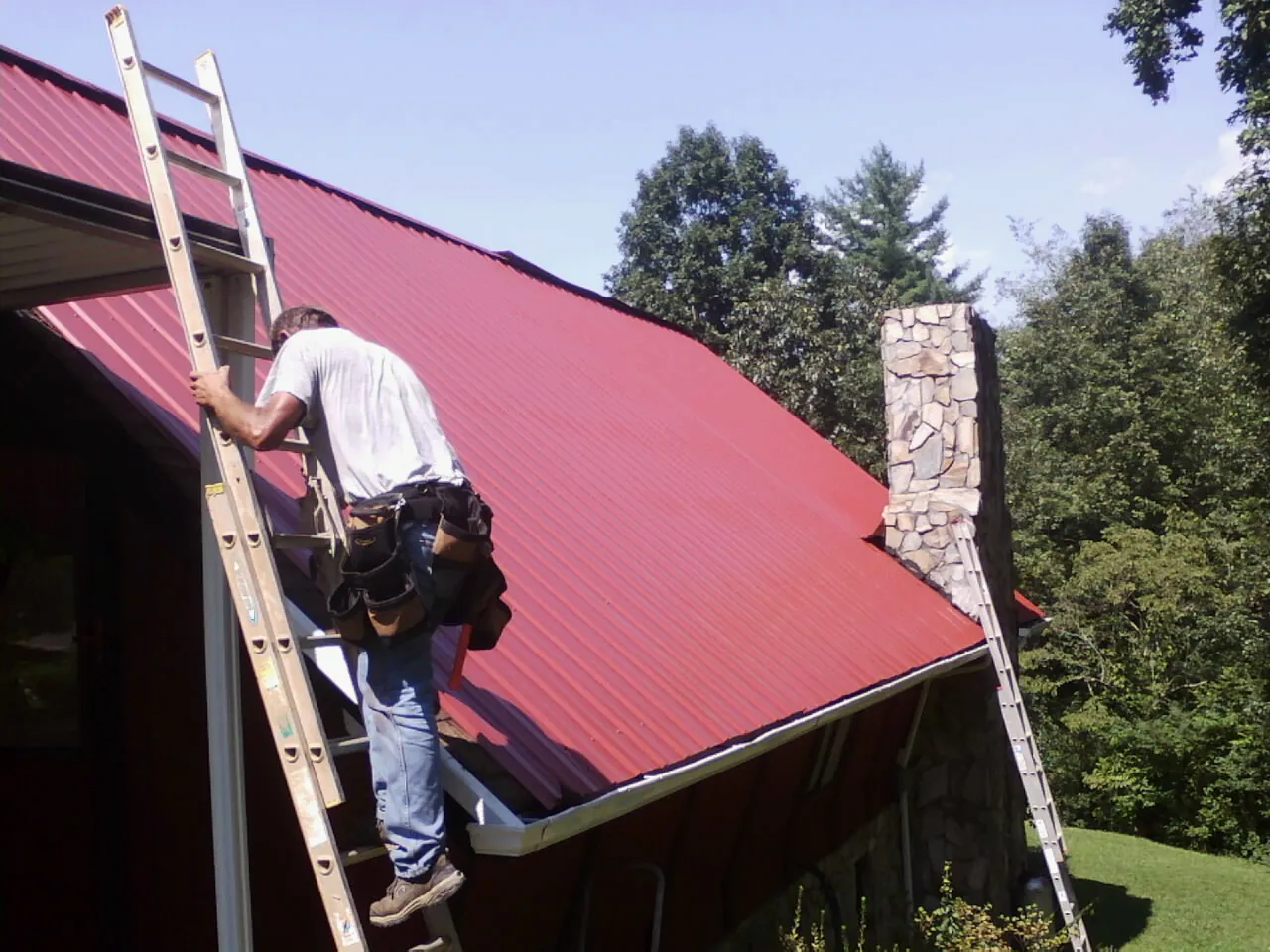 Roofer on a ladder beside a red metal roof and stone chimney — Soffit & Fascia Repair by Roofing Brothers in Secaucus